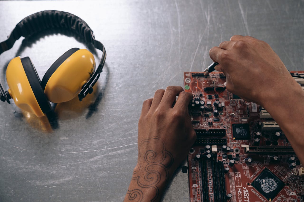 Hands repairing a motherboard on a metallic surface with yellow earmuffs beside it.